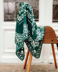 The Secret Garden 100% Silk in Vert scarf by Maison Lecomte Flament, featuring white and pink floral patterns, is draped over a wooden chair before a white paneled door with glass inserts.