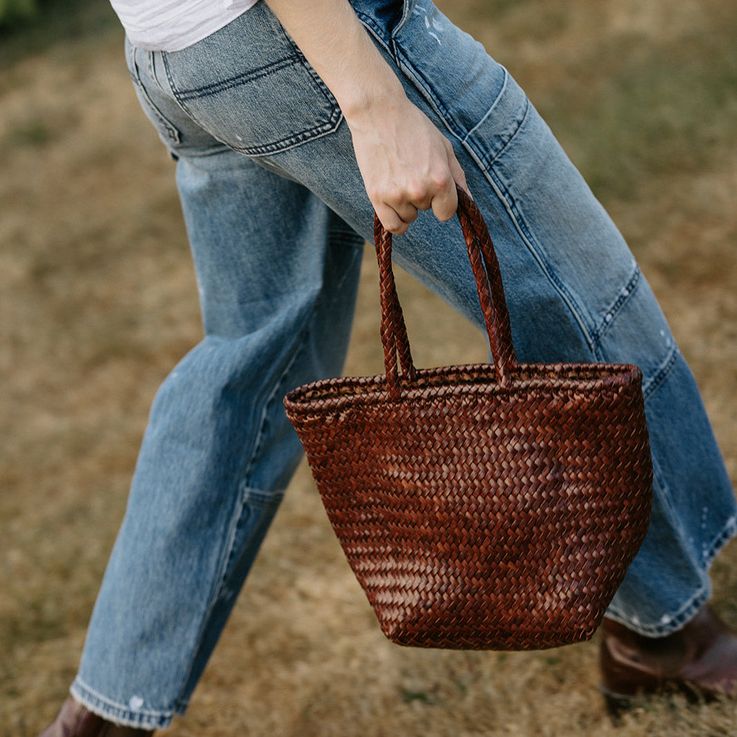 Person holding a woven brown bag outdoors on a grassy area