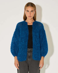 A woman with straight, light brown hair models the Maiami Mohair Big Cardigan in Royal Blue over a black shirt and gray jeans, standing against a plain white background.