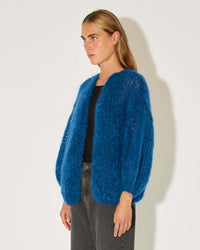 A woman with straight, light brown hair wears the Maiami Mohair Big Cardigan in Royal Blue over a black top and dark jeans, standing against a plain white background and looking to the side.