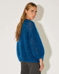 A woman with long, straight light brown hair wears the Maiami Mohair Big Cardigan in Royal Blue and dark gray pants, standing sideways and looking over her shoulder against a plain white background.