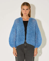 A person wears the Maiami Mohair Bomber Cardigan in Dove Blue over a black top and high-waisted dark gray jeans, standing against a plain white background and looking directly at the camera.