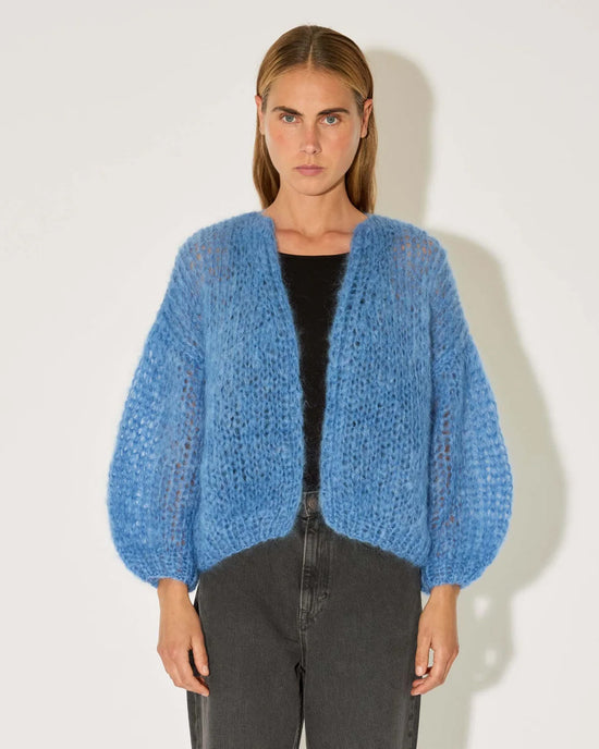 A person wears the Maiami Mohair Bomber Cardigan in Dove Blue over a black top and high-waisted dark gray jeans, standing against a plain white background and looking directly at the camera.