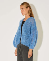 A person with long, straight hair models the Maiami Mohair Bomber Cardigan in Dove Blue over a black top and dark pants, standing against a plain white background.
