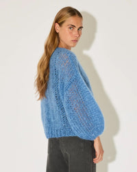 A person with long, light brown hair stands sideways, looking at the camera. They wear Maiami's oversized Mohair Bomber Cardigan in Dove Blue and dark gray jeans against a plain white background.