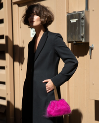 Woman in black coat with Marian Paquette Susan Solid Mohair Clutch in Fuchsia standing confidently in sunlight against a wooden door.