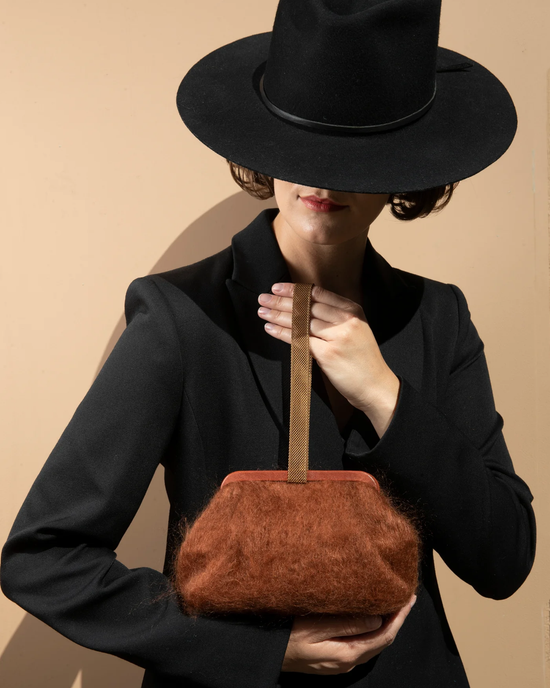 A person in a black outfit and wide-brimmed hat holding a Marian Paquette Susan Solid Mohair Clutch in Chestnut with a vintage chain handle against a beige background.