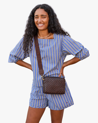 A woman smiling, wearing a blue and white striped outfit and a brown crossbody bag with an Adj Crossbody Strap in Chocolate w/ Block Ciao by Clare V., standing against a white background.