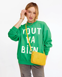 A person in a green sweatshirt with "yout bien" text, holding a Midi Sac in Dandelion Rattan with a cross-body strap by Clare V, and wearing a chunky necklace, against a