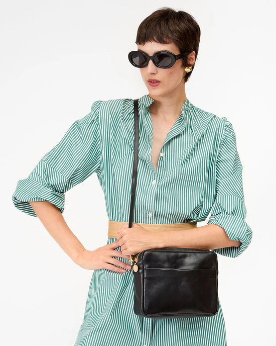 A woman in a green and white striped dress, black sunglasses, and gold earrings stands with one hand on her hip, carrying the Clare V. Mirabel in Black Rustic bag over her shoulder against a plain background.