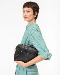 A woman with short brown hair in a green and white striped dress holds the Clare V. Mirabel in Black Rustic leather bag under her arm, standing against a plain white background and glancing over her shoulder.