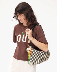 A woman with wavy brown hair wears a brown OU t-shirt and white pants, holding the Clare V. Woven Checker Moyen Messenger in Black & Cream, standing against a plain white background.