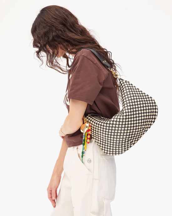 A person with wavy brown hair in a brown t-shirt and white pants stands sideways, carrying the Clare V. Woven Checker Moyen Messenger in Black & Cream adorned with colorful beaded charms.