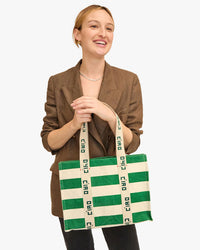 A smiling woman in a brown blazer holding a Clare V. Noemie in Palm Green & Natural Canvas Stripe tote bag.