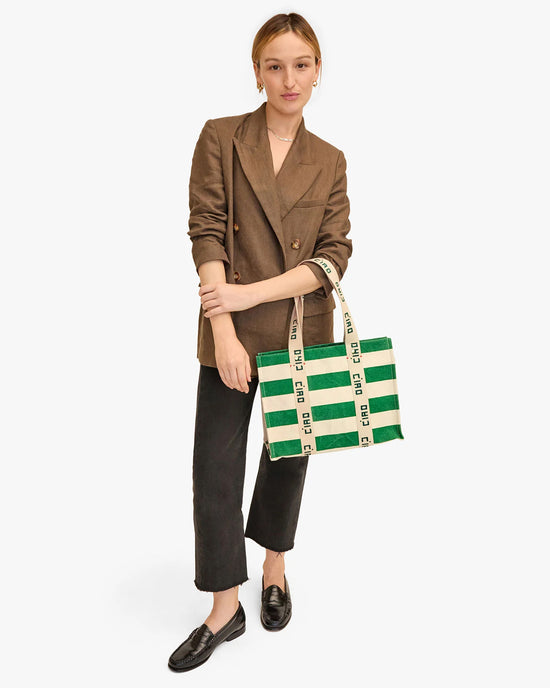 A young woman in a brown blazer and black trousers holding a Clare V. Noemie in Palm Green & Natural Canvas Stripe tote, standing against a white background.