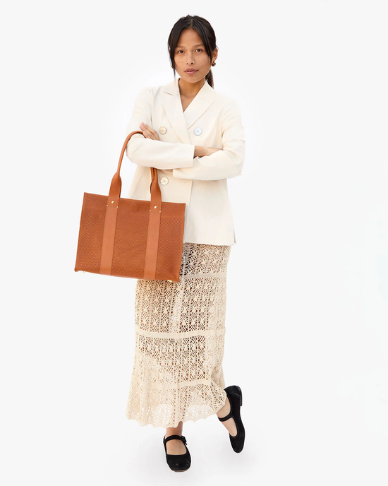 A woman standing against a white background, wearing a light blazer, a lacy skirt, holding a Clare V. Noemie in Cuoio Perf perforated leather tote bag, and black shoes.