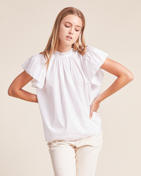 Woman posing in a white Trovata Birds of Paradis Carla Highneck Shirt with ruffle sleeves and beige pants.