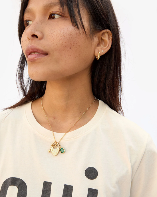 Woman wearing a necklace with a Clare V. Padlock Locket in Vintage Gold charm and stud earring, with a cream-colored top featuring the word "oui".