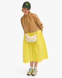 Woman in a brown blouse and yellow skirt, wearing sneakers, a cap, and carrying a Clare V. Petit Moyen Messenger - Woven Checker in Cream, standing against a white background.