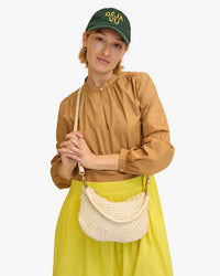 A woman wearing a yellow cap labeled "deja vu," a tan blouse, and a bright yellow skirt, holding a Clare V. Petit Moyen Messenger - Woven Checker in Cream, standing against a white background.