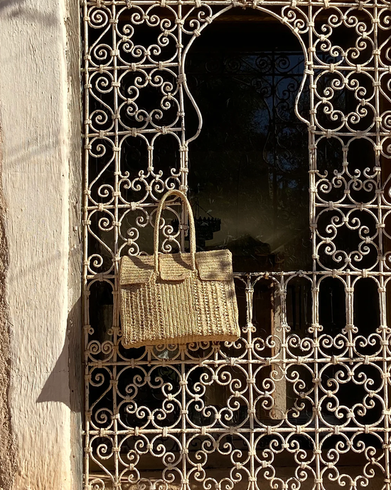 A Maison N.H Paris Dahlia Mini Bag in Natural, a raffia flap bag, hanging on an ornate metal grille covering an arched window, with sunlight casting shadows on a textured wall.