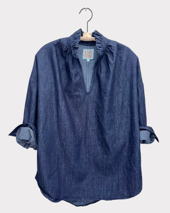 A Penelope - Denim in Indigo blouse with ruffle collar and tie-up sleeves hanging on a wooden hanger against a white background. (Brand Name: A Shirt Thing)