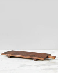 The Bordeaux Footed Tray by etúHOME, a rectangular wooden cheese board with a handle, resting on a marble surface against a gray background, embodies the ideal blend of rustic modern kitchen aesthetics.