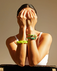 A person covering their face with their hands, wearing brightly colored bracelets, including a distinctive Clare V. Resin Link Bracelet in Neon Yellow.