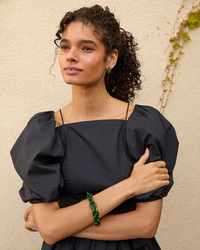 A woman with curly hair wearing a dark puff-sleeve dress and green accessories, including a Clare V. Fern resin link bracelet, poses with her arms crossed.