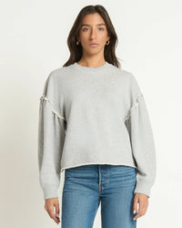 A woman stands against a plain background wearing the Shane Sweatshirt in Heather Grey by Nation LTD and blue jeans, looking directly at the camera with a neutral expression.