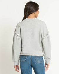 A woman with long brown hair, wearing the Shane Sweatshirt in Heather Grey by Nation LTD with ruffle sleeve details and blue jeans, stands facing away from the camera against a plain white background.