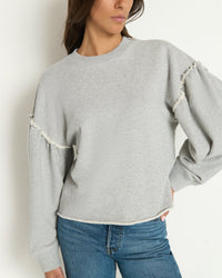 A woman wearing the Shane Sweatshirt in Heather Grey by Nation LTD, featuring a structured crewneck and ruffled sleeves, pairs it with blue jeans and stands with one hand on her hip against a plain white background.