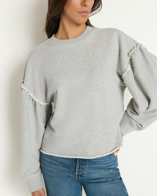 A woman wearing the Shane Sweatshirt in Heather Grey by Nation LTD, featuring a structured crewneck and ruffled sleeves, pairs it with blue jeans and stands with one hand on her hip against a plain white background.