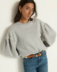 A woman with long brown hair wears the Nation LTD Shane Sweatshirt in Heather Grey, featuring a structured crewneck and puffed sleeves, paired with blue jeans and a brown belt. She stands against a light wall, gazing confidently at the camera.
