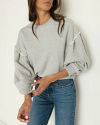 A woman wears the Shane Sweatshirt in Heather Grey by Nation LTD, featuring puffed sleeves and frayed seams, styled with high-waisted blue jeans against a plain white wall.