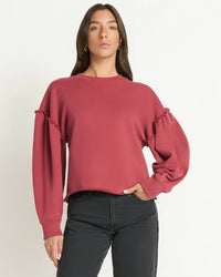 A woman in the Nation LTD Shane Sweatshirt in Russet—featuring long sleeves, a loose fit, ruched sleeve details—and black jeans stands against a plain light background.