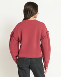 A woman with long dark hair, seen from behind against a plain light background, wears the Nation LTD Shane Sweatshirt in Russet with ruffle sleeve details and black jeans.