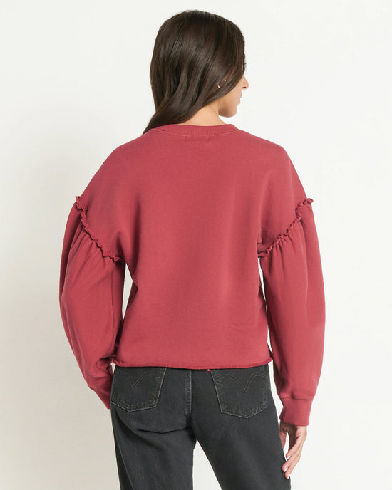 A woman with long dark hair, seen from behind against a plain light background, wears the Nation LTD Shane Sweatshirt in Russet with ruffle sleeve details and black jeans.