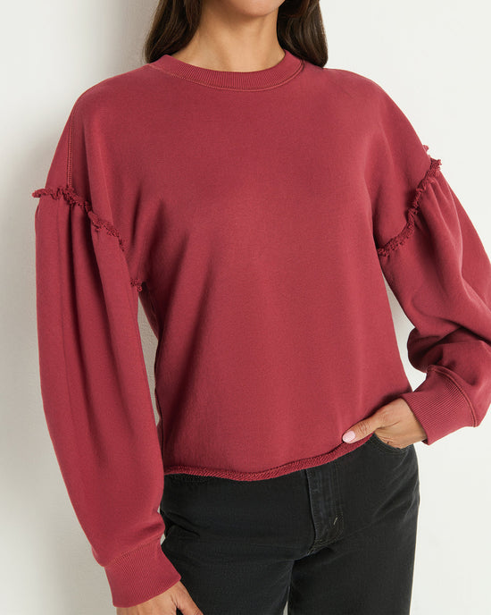 A woman wears the Nation LTD Shane Sweatshirt in Russet—a long-sleeve, loose-fitting vintage top with frayed sleeves and a structured crewneck—paired with black pants, standing against a plain light background.