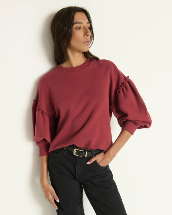 A woman wearing the Shane Sweatshirt in Russet by Nation LTD and black pants stands against a plain white wall, looking to the side with one hand on her hip.
