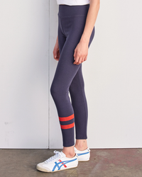 A person standing in profile view showcasing Sundry's Striped Leggings in Navy, made from a cotton/modal blend with orange stripes at the calf and wearing white sneakers.