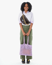 Woman standing and smiling, carrying a Clare V Sandy in Lilac Woven Bag and wearing a lilac shirt with green trousers.
