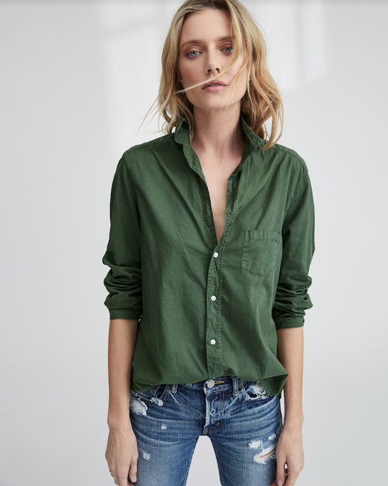 A woman with wavy blonde hair wears the Frank & Eileen Eileen Relaxed Button Up Shirt in army green, styled with blue distressed jeans, standing against a light, neutral background.