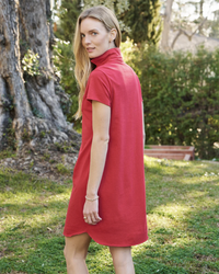 A woman in a Frank & Eileen 100% Cotton Heritage Jersey, Lauren S/S Polo Dress in Double Decker Red with a high neck stands in a garden, looking over her shoulder at the camera.