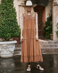 Woman in a tiered terracotta the Great Soleil Dress in Canyon with floral embroidery and wide-brimmed hat standing in front of a building with manicured shrubbery.