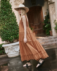 A woman in a flowing brown The Soleil Dress in Canyon by the Great with a hand-crocheted chest panel and a wide-brimmed hat walking past a building with an ornate door and a manicured bush.
