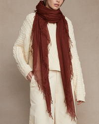 A person in a cream cable-knit sweater and beige pants accessorized with a long, rust-colored Chan Luu Cashmere & Silk Scarf in Fired Brick with fringed trims.