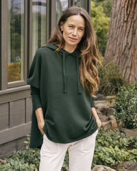 Woman in a Frank & Eileen Kane Capelet Hoodie in Evergreen and white pants standing outdoors.