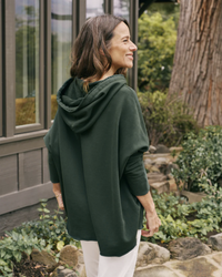 Woman wearing a Frank & Eileen Kane Capelet Hoodie in Evergreen and white pants standing outside near a building and vegetation.