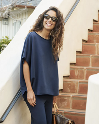 A woman smiling while leaning against a stair railing, wearing a casual Frank & Eileen Olive Capelet in Atlantic top, pants, and sunglasses, with a handbag by her side.